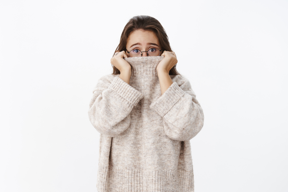 Indoor shot of insecure worried young cute woman in glasses with brown hair pulling collar of sweater on nose and frowning nervously as looking scared at camera, posing anxious over gray background - Image( Cookie Studio)s