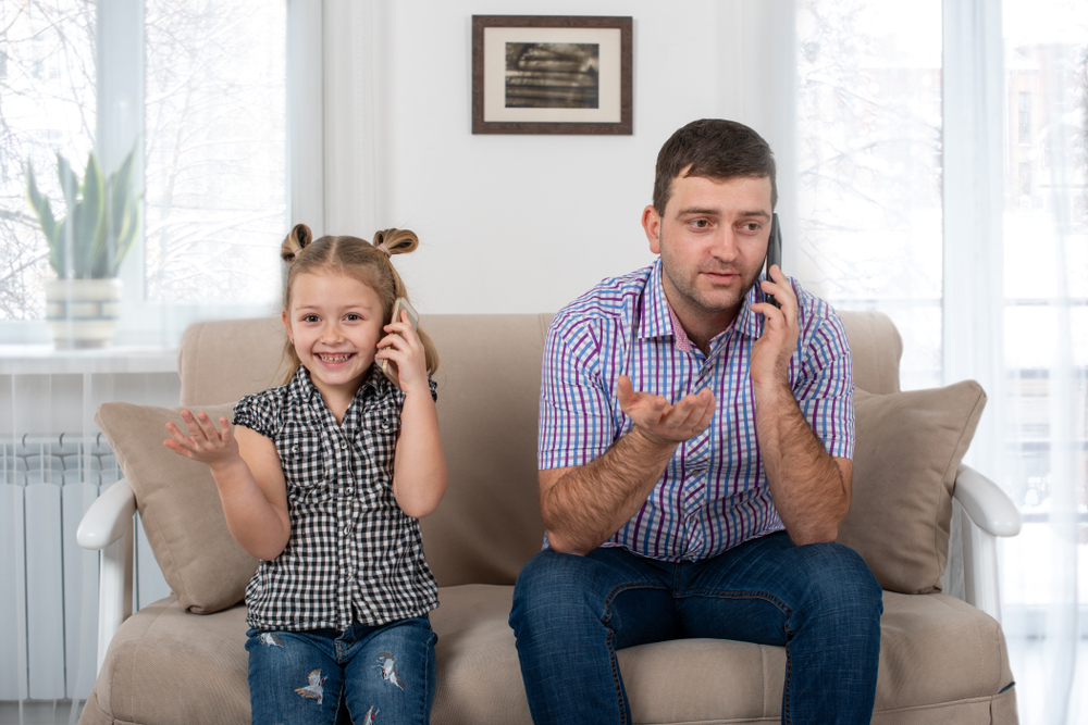 Studio shot of a daughter and dad sitting on the sofa at home and talking on the phone the same way(Rozochka)s