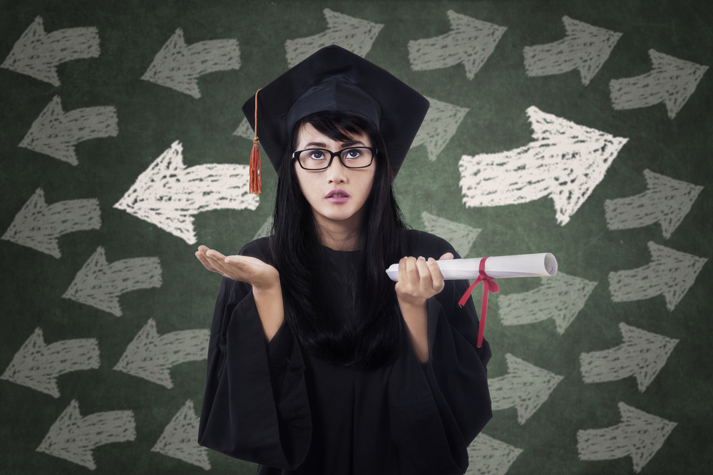 Confused young female in graduation gown with arrow symbol on the blackboard - Image( Creativa Images)s