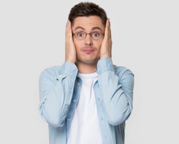 Funny worried man in glasses jean shirt hold head in hands looks concerned pose studio grey wall(fizkes)S