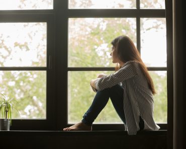 Thoughtful girl sitting on sill embracing knees looking at window(fizkes)s