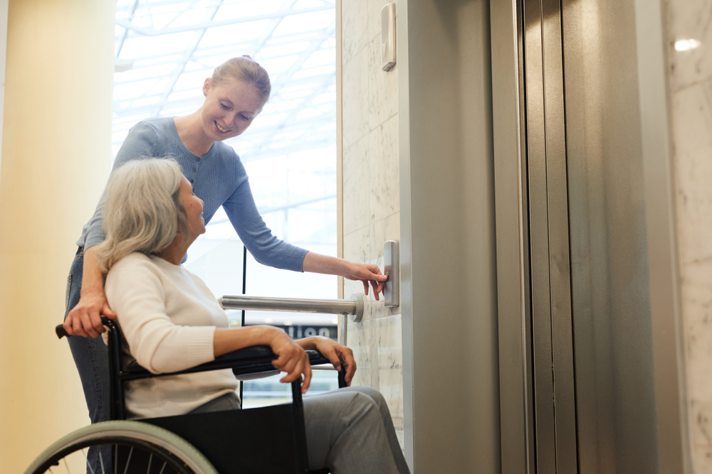 Young woman pushing the button of elevator and smiling to senior disabled woman while she sitting in wheelchair